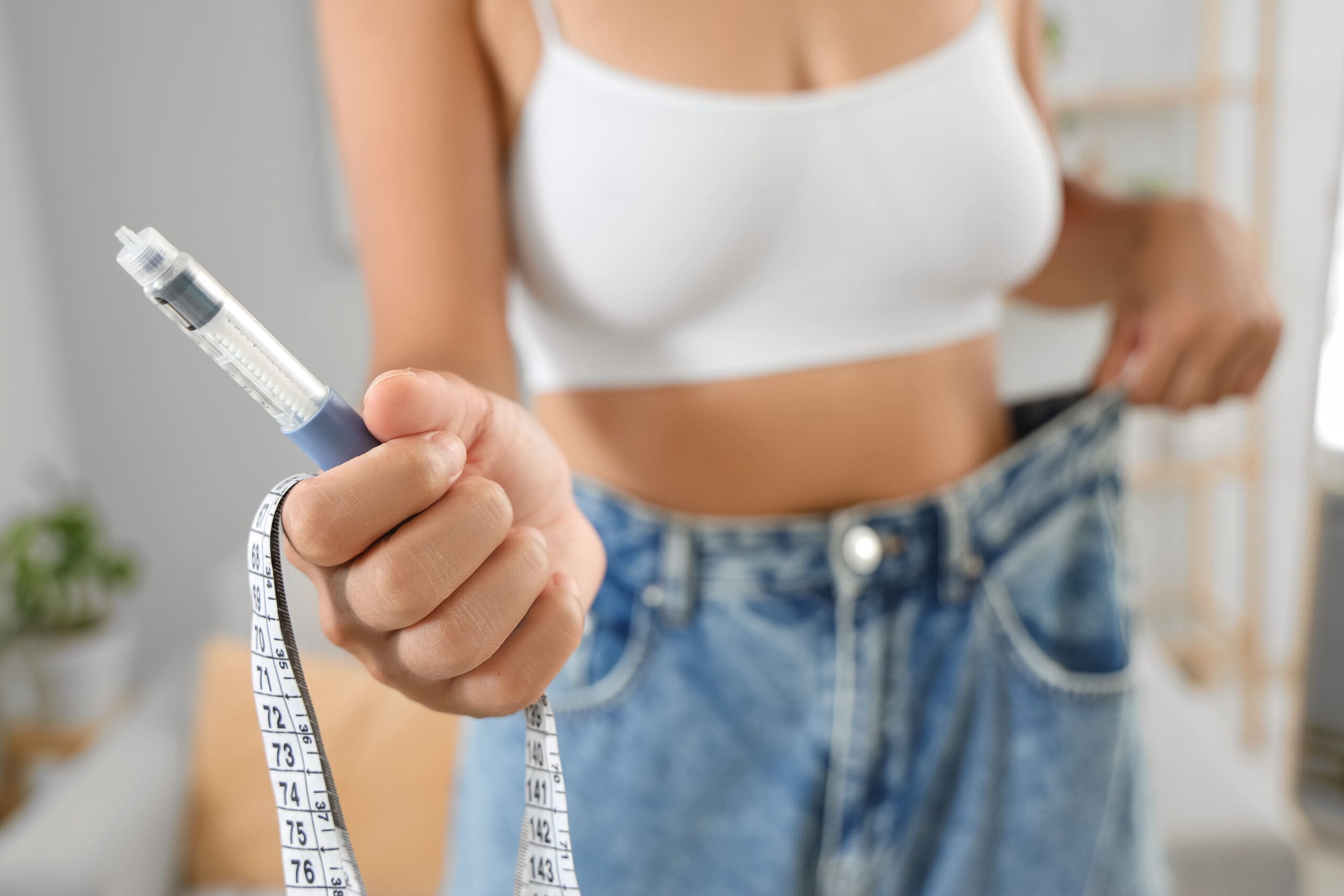 Woman in loose jeans with injection and tape measure at home, closeup. Weight loss concept